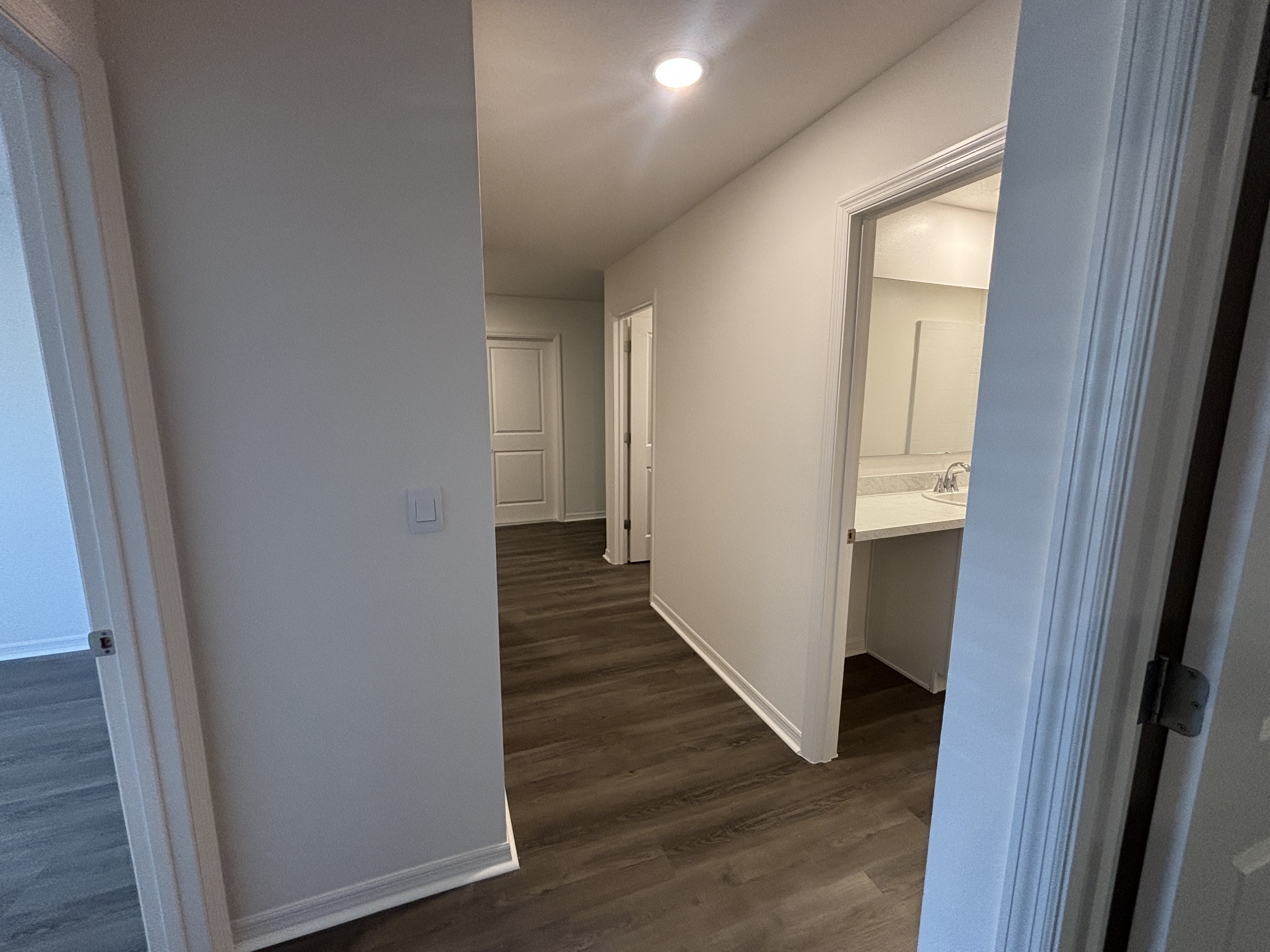 A modern hallway with dark wood flooring and white walls, leading to a bathroom with a visible sink.