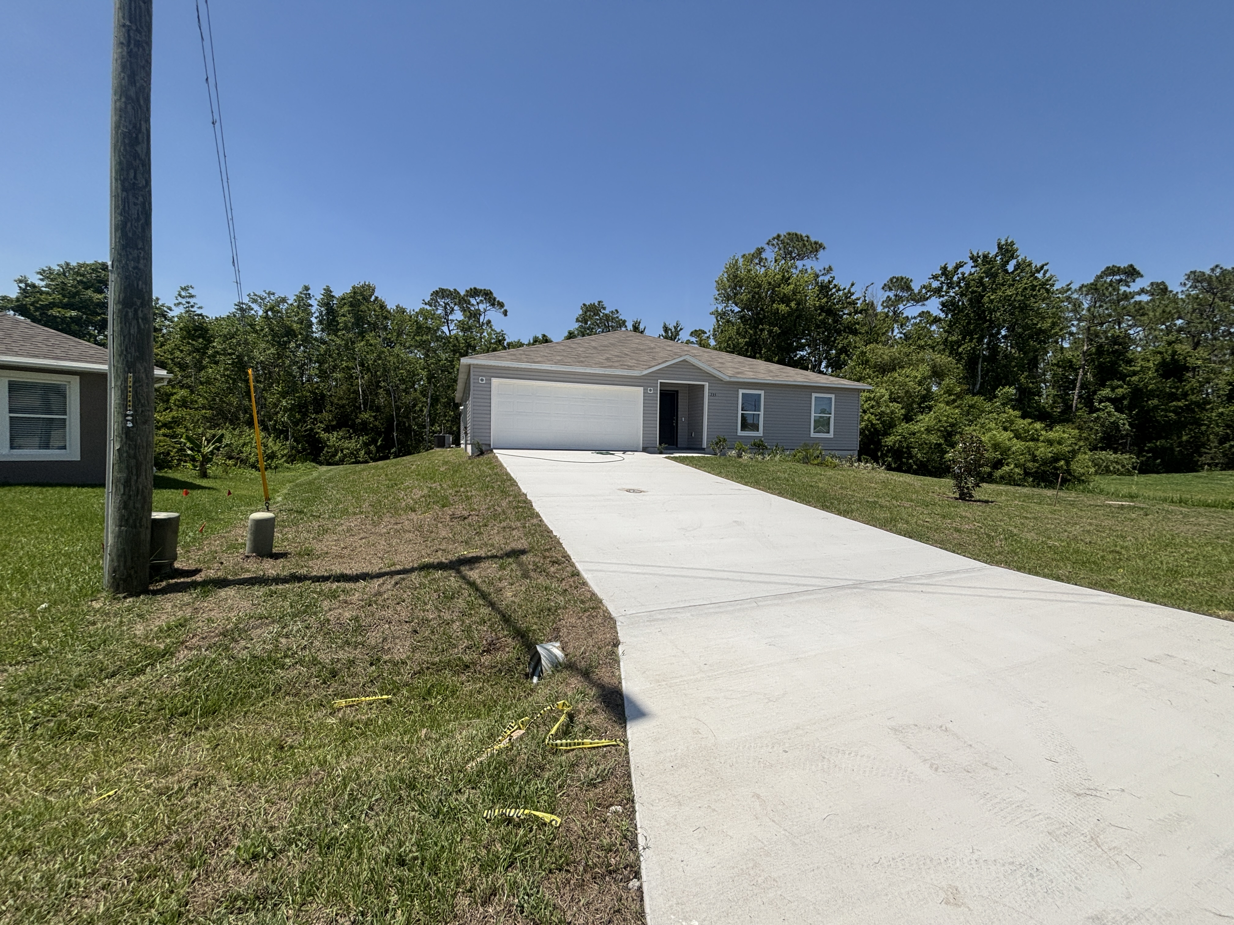 Single-story, gray house with a large concrete driveway surrounded by lush greenery on a clear sunny day.