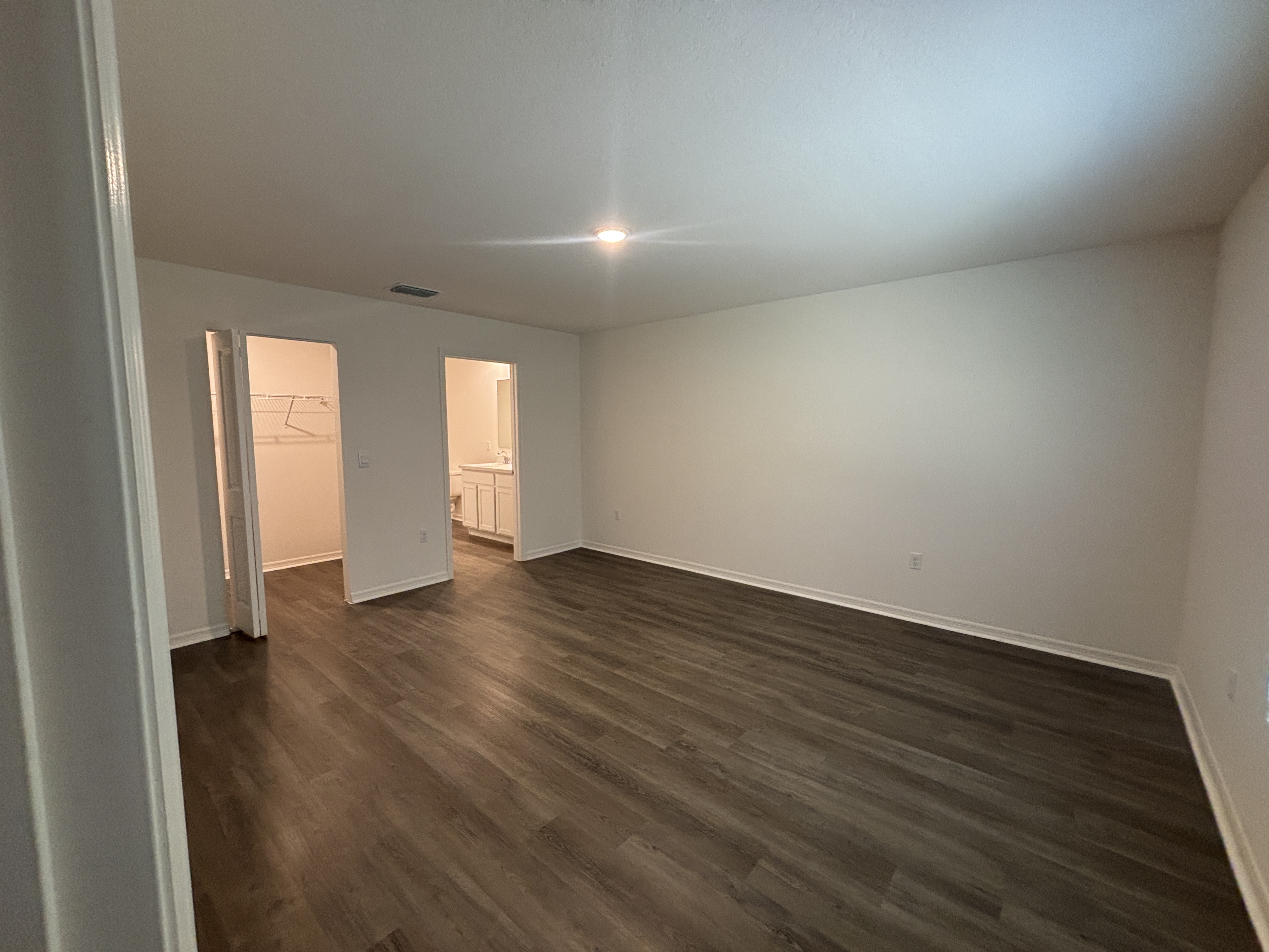 Spacious empty room with hardwood flooring, white walls, and doorway leading to a closet and bathroom with light fixtures.